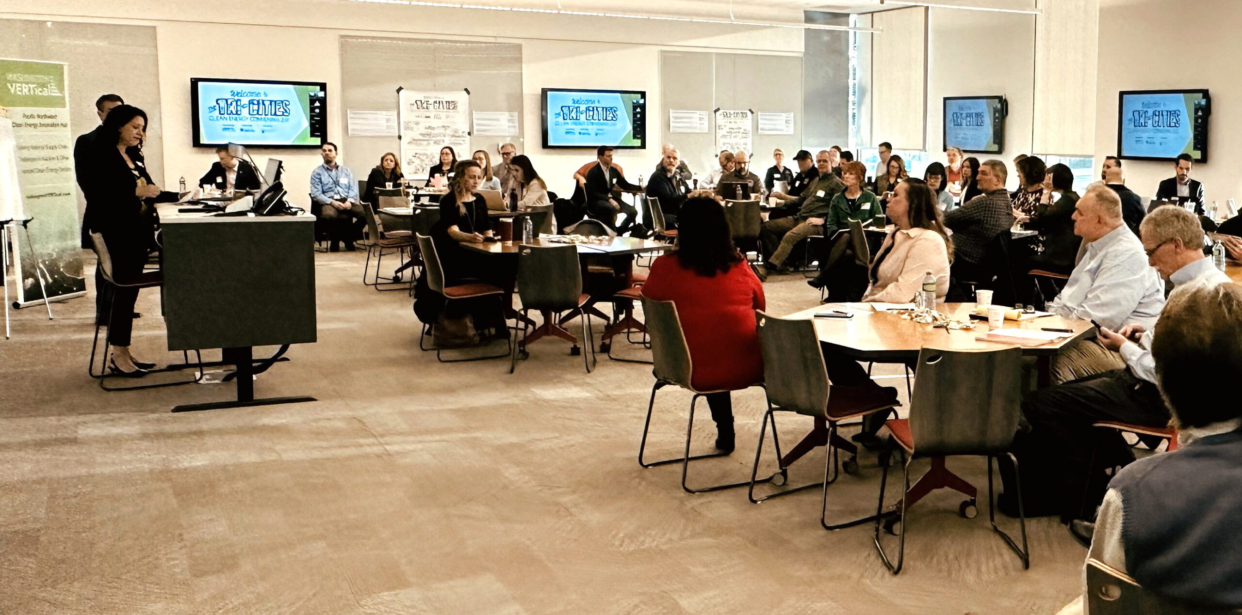 Room full of people sitting at tables with their attention on a woman speaking while standing at a podium.