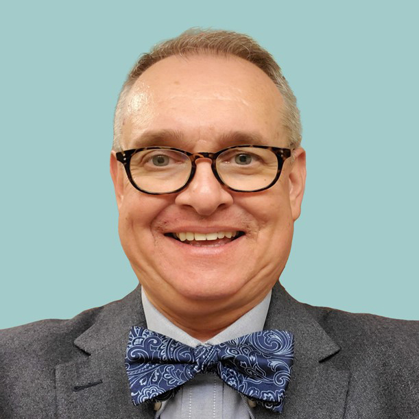 Portrait of John Gonsky wearing glasses, a blue bow tie, light gray shirt and gray jacket on a light blue background.