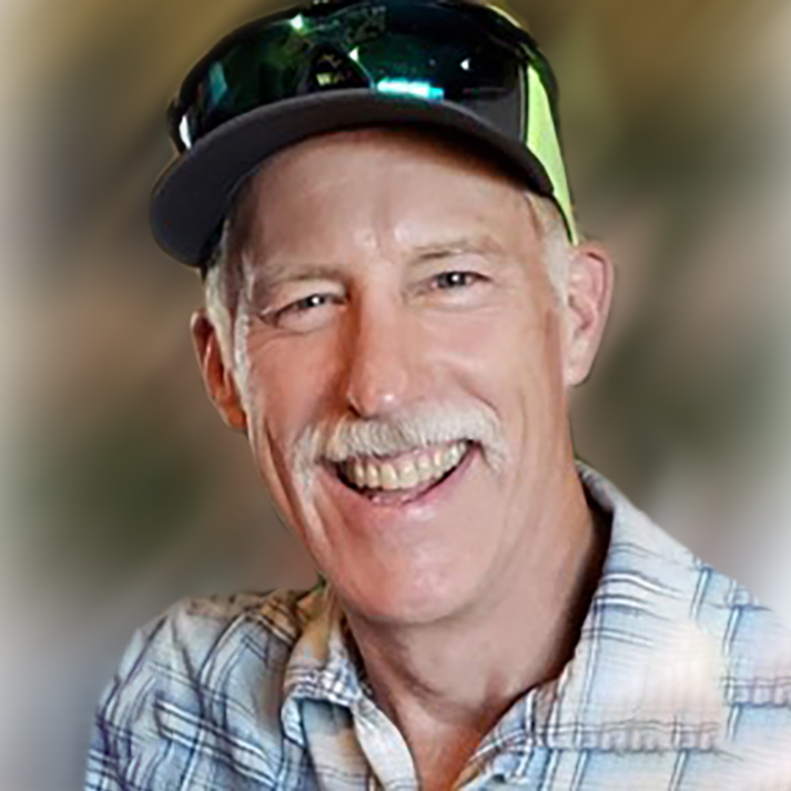 Portrait of Steve Anderson wearing a blue plaid shirt, black and green baseball cap with sunglasses sitting on the hat brim.